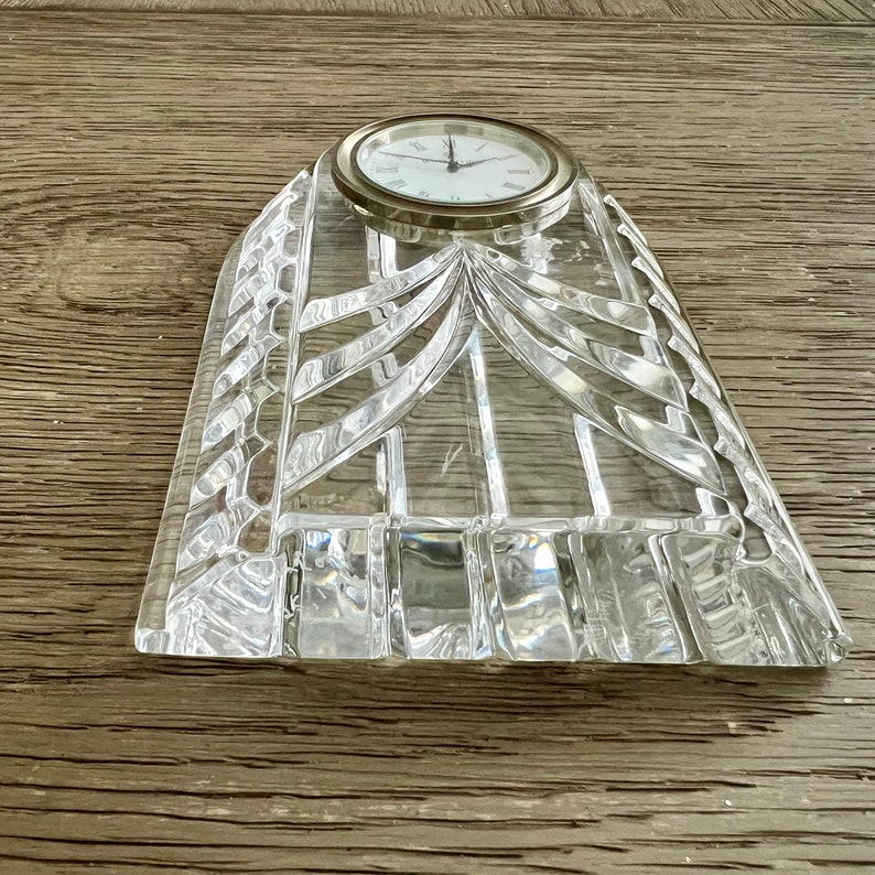 May include: A clear crystal clock with a silver face and black hands. The clock is on a clear crystal base with a geometric design.