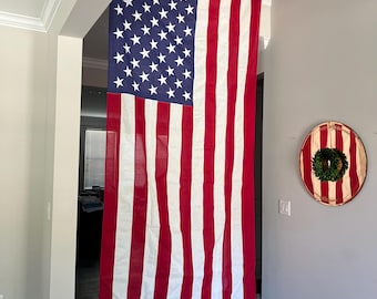 May include: A large American flag with red and white stripes and a blue field of white stars hangs indoors. The flag is displayed in a doorway. A round, decorative piece with red and white stripes and a green wreath hangs on the wall.