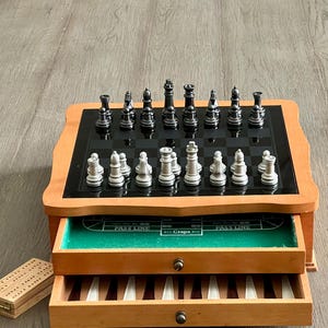 Puede incluir: Una caja de juegos de madera con un tablero de ajedrez blanco y negro, un tablero de dados de fieltro verde y dos cajones que contienen fichas de dominó y clavijas de cribbage. Las piezas de ajedrez son plateadas y negras.