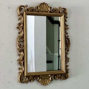 May include: A rectangular gold-colored mirror with an ornate, detailed frame. The frame features scrolling floral and shell-like designs. The mirror is hanging on a white wall, reflecting a glimpse of a staircase.