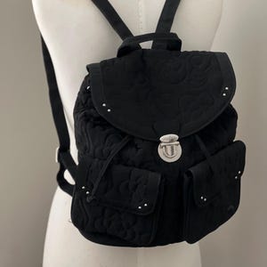 Può includere: Zaino nero trapuntato con motivo floreale. Presenta due tasche frontali, borchie argentate e chiusura con coulisse. La patta superiore si chiude con un fermaglio argentato. Spallacci regolabili.