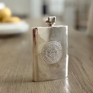 Puede incluir: Una petaca rectangular plateada con tapón de rosca. La petaca presenta un emblema circular detallado en la parte delantera. La petaca está hecha de un metal brillante y reflectante y está sobre una superficie de madera.