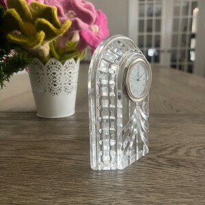 May include: A clear crystal clock with a white face and black Roman numeral hour markers. The clock is in the shape of an arch and has a decorative design.