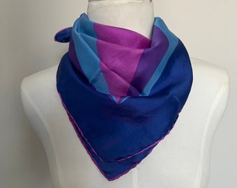 May include: A blue silk scarf with a geometric pattern in shades of purple and blue. The scarf is folded into a triangle shape and is displayed on a white mannequin.