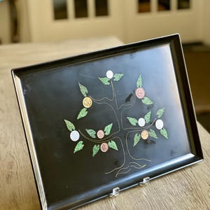 Puede incluir: Bandeja rectangular negra con un diseño de árbol dorado. El árbol tiene hojas de purpurina verde y varias monedas como fruta. La bandeja se exhibe sobre una superficie de madera clara.