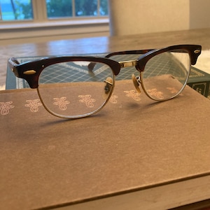 Könnte beinhalten: Schildpattbrille mit goldenen Akzenten und klaren Gläsern. Die Brille liegt auf einem braunen Buch.