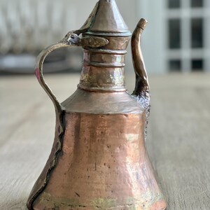May include: A vintage copper teapot with a hammered finish and a pointed lid. The teapot has a curved spout and a handle with a decorative design.