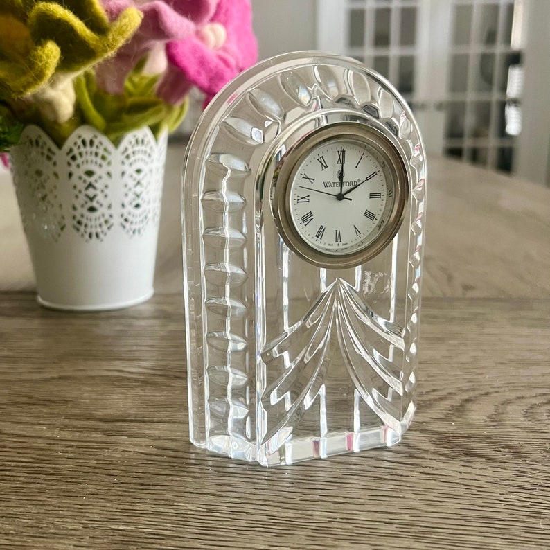 May include: A Waterford crystal clock with a silver face and Roman numeral hour markers. The clock is in a clear crystal frame with a decorative design.