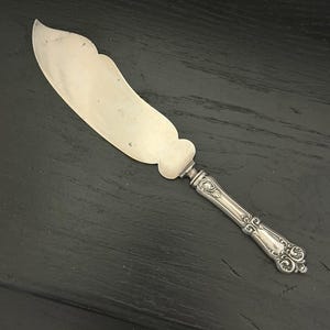 May include: Silver butter knife with a curved blade and ornate handle. The knife has a decorative design on the handle and a wide, slightly curved blade. The handle features detailed scrollwork and a rounded end.