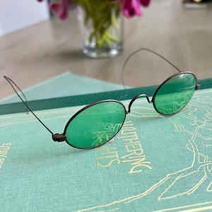 May include: Vintage round spectacles with green lenses and a dark metal frame. The glasses are placed on a book with gold lettering. The lenses reflect the surrounding environment.