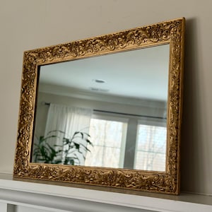 Pode incluir: Espelho retangular com moldura dourada ornamentada. A moldura apresenta detalhes de rolagem e um tom dourado quente. O espelho reflete uma sala com uma janela e uma planta, adicionando profundidade e dimensão à imagem.