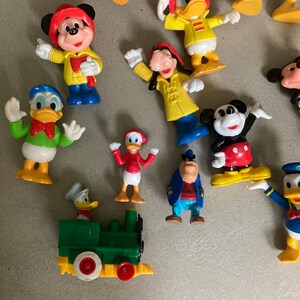 May include: A collection of vintage Disney character figurines, including Mickey Mouse, Donald Duck, Goofy, and Pluto. The figurines are made of plastic and are painted in bright colors. They are all standing on a white surface.