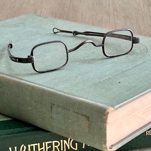 May include: A pair of antique, silver-toned, folding reading glasses with square lenses. The glasses are resting on a green book with the title "Wuthering Heights" visible.