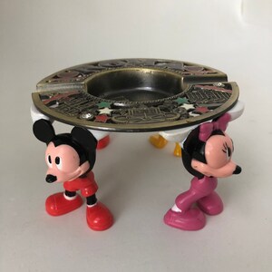 K&ouml;nnte beinhalten: Ein vintage Aschenbecher aus Metall mit einem Mickey Mouse und Minnie Mouse Design. Der Aschenbecher ist mit einem goldfarbenen Design einer Stadtlandschaft und Sternen verziert. Der Aschenbecher wird von vier Plastikfiguren von Mickey Mouse und Minnie Mouse getragen.