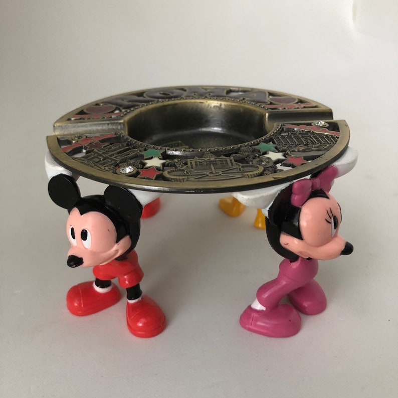 K&ouml;nnte beinhalten: Ein vintage Aschenbecher aus Metall mit einem Mickey Mouse und Minnie Mouse Design. Der Aschenbecher ist mit einem goldfarbenen Design einer Stadtlandschaft und Sternen verziert. Der Aschenbecher wird von vier Plastikfiguren von Mickey Mouse und Minnie Mouse getragen.