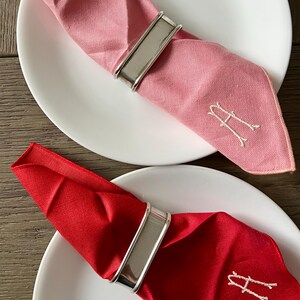 May include: Two white plates with pink and red napkins. Each napkin is held by a silver napkin ring. The pink napkin has a white embroidered letter "A". The red napkin also has a white embroidered letter "A".