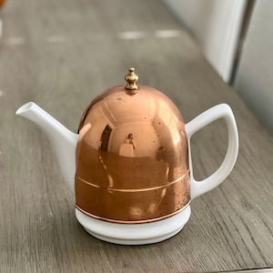 Könnte beinhalten: Eine Kupfer- und weiße Teekanne mit einem abgerundeten Kupferdeckel und einem kleinen goldenen Knauf. Die Teekanne hat einen weißen Ausguss und Henkel. Die Kupferoberfläche reflektiert die Umgebung. Die Teekanne steht auf einer Holzoberfläche.
