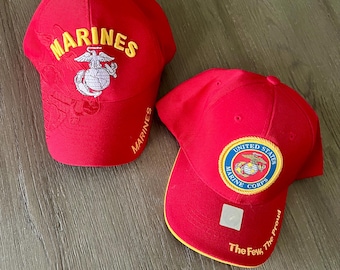 May include: Two red baseball caps with "MARINES" embroidered in yellow. One cap features the Marine Corps emblem and the other has a patch with the Marine Corps emblem and the text "The Few, The Proud".
