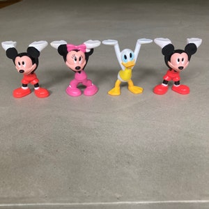 K&ouml;nnte beinhalten: F&uuml;nf kleine Plastikfiguren von Disney-Charakteren. Die Figuren sind Mickey Mouse, Minnie Mouse, Donald Duck, Goofy und Pluto. Die Figuren stehen alle in aufrechter Position mit ausgestreckten Armen.