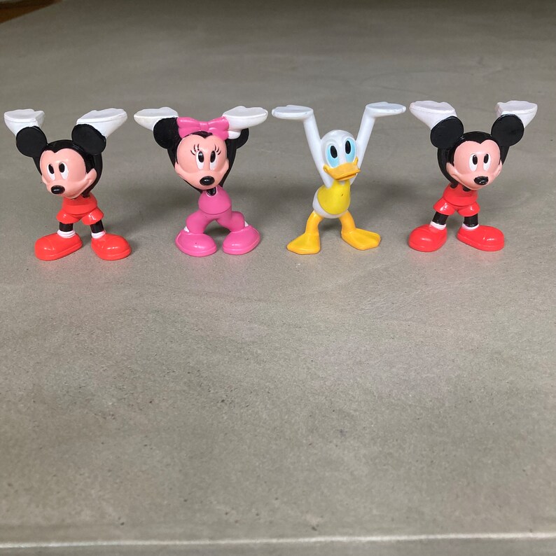 K&ouml;nnte beinhalten: F&uuml;nf kleine Plastikfiguren von Disney-Charakteren. Die Figuren sind Mickey Mouse, Minnie Mouse, Donald Duck, Goofy und Pluto. Die Figuren stehen alle in aufrechter Position mit ausgestreckten Armen.