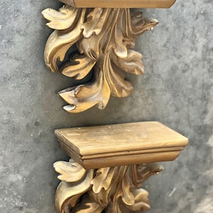 Pode incluir: Duas prateleiras de parede douradas e ornamentadas. Cada prateleira tem um tampo retangular com uma borda decorativa, sustentada por um design detalhado em forma de folha. As prateleiras são montadas em uma parede cinza texturizada.