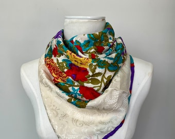 May include: A white silk scarf with a floral pattern in shades of red, yellow, blue, and green. The scarf has a paisley design on the bottom half and a purple trim.