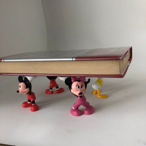 K&ouml;nnte beinhalten: Vier bunte Plastikfiguren, die ein Buch hochhalten. Die Figuren sind Mickey Mouse, Minnie Mouse, Donald Duck und Goofy. Das Buch ist rot und hat einen wei&szlig;en R&uuml;cken.