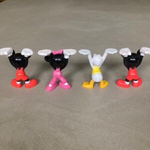 K&ouml;nnte beinhalten: Vier kleine Plastikfiguren von Comicfiguren. Die Figuren sind Mickey Mouse, Minnie Mouse, Donald Duck und Goofy. Sie tragen alle verschiedene farbige Outfits und sind mit erhobenen Armen &uuml;ber ihren K&ouml;pfen posiert.