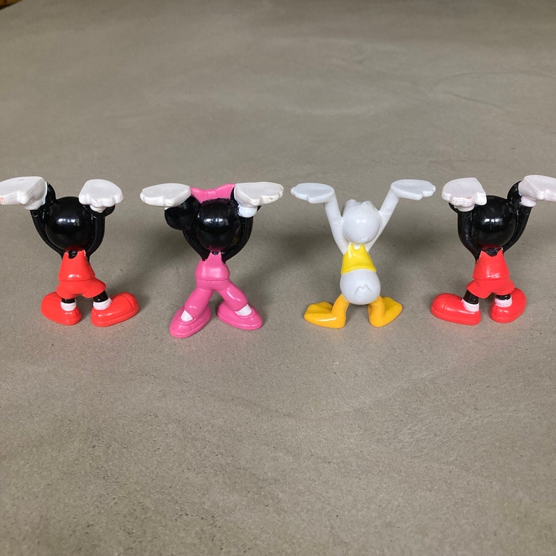 K&ouml;nnte beinhalten: Vier kleine Plastikfiguren von Comicfiguren. Die Figuren sind Mickey Mouse, Minnie Mouse, Donald Duck und Goofy. Sie tragen alle verschiedene farbige Outfits und sind mit erhobenen Armen &uuml;ber ihren K&ouml;pfen posiert.