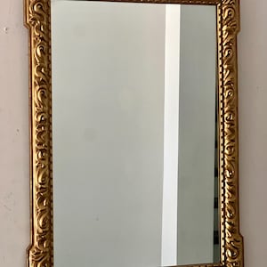 Può includere: Specchio rettangolare con cornice dorata e ornata. La cornice presenta intagli dettagliati. Lo specchio riflette una parete bianca e una colonna verticale bianca. Un elemento classico e decorativo per l'arredamento.