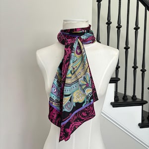 May include: A colorful silk scarf with a paisley design. The scarf features a black background with vibrant accents of pink, teal, yellow, and purple. The scarf is draped over a mannequin.