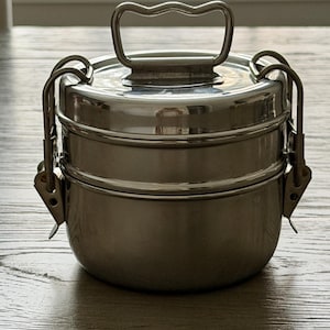 May include: A two-tiered stainless steel food container with a polished finish. The container features a round shape, a top lid with a central arched handle, and two side locking latches that secure the stacked compartments together firmly.