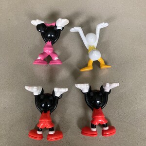 K&ouml;nnte beinhalten: Vier kleine Plastikfiguren von Comicfiguren. Zwei sind Mickey Mouse, eine ist Minnie Mouse und eine ist Donald Duck. Die Figuren stehen alle in aufrechter Position mit erhobenen Armen &uuml;ber dem Kopf. Die Mickey Mouse Figuren tragen rote und schwarze Shorts, die Minnie Mouse Figur tr&auml;gt rosa Shorts und die Donald Duck Figur tr&auml;gt gelbe Shorts.