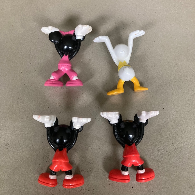 K&ouml;nnte beinhalten: Vier kleine Plastikfiguren von Comicfiguren. Zwei sind Mickey Mouse, eine ist Minnie Mouse und eine ist Donald Duck. Die Figuren stehen alle in aufrechter Position mit erhobenen Armen &uuml;ber dem Kopf. Die Mickey Mouse Figuren tragen rote und schwarze Shorts, die Minnie Mouse Figur tr&auml;gt rosa Shorts und die Donald Duck Figur tr&auml;gt gelbe Shorts.