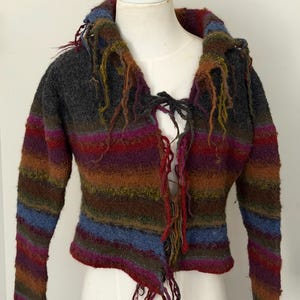 May include: A long-sleeved, cropped knit sweater featuring horizontal stripes in shades of charcoal gray, deep purple, rust orange, and muted blue. The garment includes a wide collar and a front opening secured with thin, dangling yarn ties.