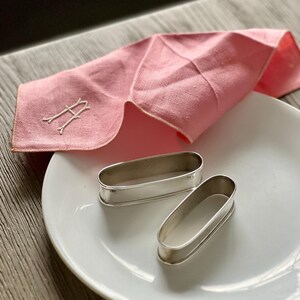 May include: A pink linen handkerchief with an embroidered "A" and two silver napkin rings on a white plate. The handkerchief is folded, showing its texture and the delicate embroidery. The napkin rings are oval-shaped.