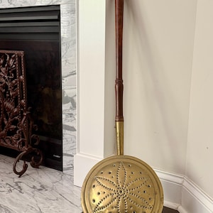 May include: Antique brass fireplace bed warmer with a long wooden handle. The round warming pan features a starburst pattern of small holes. The handle is dark brown with a brass connector.
