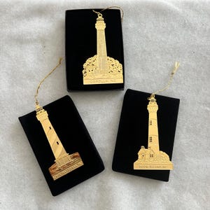 Op de afbeelding: Drie gouden lichtbaken ornamenten in zwarte fluwelen doosjes. De ornamenten zijn gedetailleerd met de namen van de vuurtorens: Corolla, NC, Cape Hatteras, NC en Bodie Island, NC.