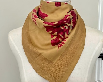 May include: A square scarf with a tan background and a burgundy floral pattern. The scarf has a patterned border and is displayed on a mannequin. The scarf is made of a soft, flowing fabric.