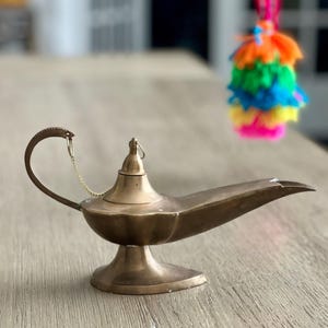 May include: A brass-colored genie lamp with a curved handle and a long spout. The lamp sits on a round base and has a small lid. A colorful tassel hangs in the background.