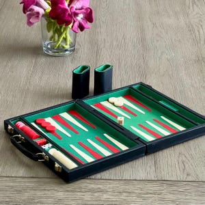 May include: A black leather travel backgammon set with a green felt playing surface, red and white playing pieces, and a white die. The set is open and ready to play.