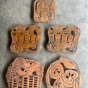 Puede incluir: Cinco posavasos de metal de color cobre con diseños de manzanas y frutas. Los posavasos tienen forma de frasco, carro, cesta, círculo y círculo con hojas.