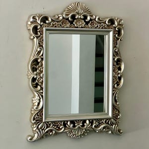 May include: Silver-colored rectangular mirror with an ornate, floral-patterned frame. The frame has a detailed, sculpted design with flowers and scrollwork. The mirror is mounted on a white wall, reflecting the room.