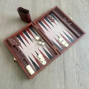 Puede incluir: Un juego de backgammon de madera marrón con un tablero de juego blanco y negro, dados y fichas. El juego está abierto y listo para jugar.