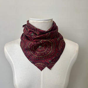 May include: A red paisley patterned bandana with green and black accents. The bandana is folded into a triangle shape and is displayed on a white mannequin.