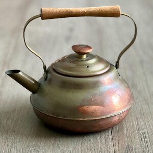 May include: A vintage copper tea kettle with a wooden handle. The kettle has a tarnished copper finish and a brass spout.