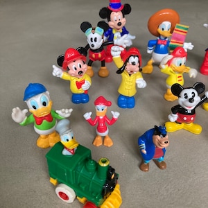 May include: A collection of 14 colorful plastic figurines of Disney characters, including Mickey Mouse, Donald Duck, Goofy, and Pluto. The figurines are all standing on a light brown surface. There is also a green toy train with a yellow smokestack.