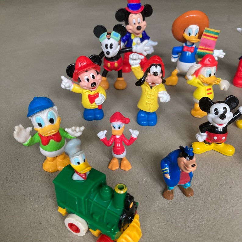 May include: A collection of 14 colorful plastic figurines of Disney characters, including Mickey Mouse, Donald Duck, Goofy, and Pluto. The figurines are all standing on a light brown surface. There is also a green toy train with a yellow smokestack.