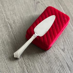 Op de afbeelding: Een zilveren taartschep met een decoratief handvat rust op een rode, gewatteerde fluwelen doos. De schep heeft een breed, plat lemmet en een gedetailleerd handvat. De achtergrond is een grijs houten oppervlak.