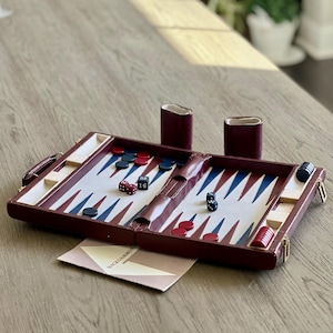 Puede incluir: Un juego de backgammon burdeos con fichas rojas, azules y blancas. El tablero está abierto, mostrando dados y una partida en curso. Se ven dos cubiletes burdeos. Una tarjeta con la palabra "BACKGAMMON" está en el tablero.
